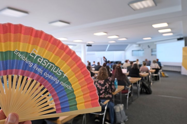 Ein Regenbogenfächer vor einem Versammlungsraum mit der Aufschrift: Erasmus+ Enriching lives, opening minds.