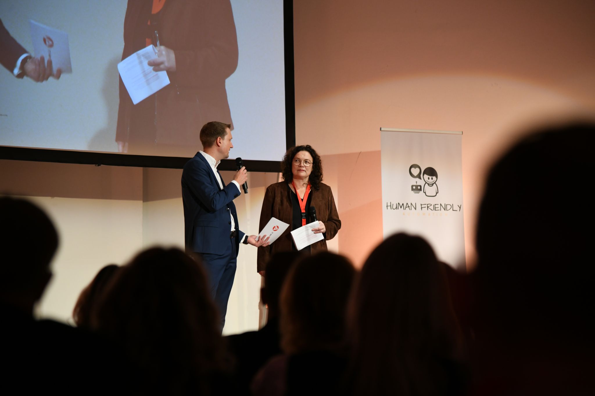 Zwei Konferenzsprecher, ein Mann und eine Frau, präsentieren auf einer Bühne vor Publikum. Im Hintergrund ein Banner mit einem Roboter, Menschen und dem Schriftzug „Human Friendly“.
