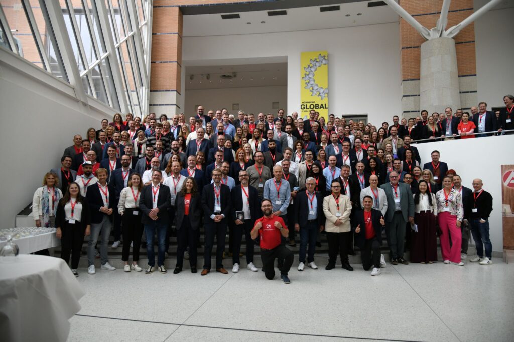 Großes Gruppenfoto von Teilnehmenden einer Konferenz. Die Personen stehen auf einer Treppe in einem modernen Gebäude und tragen Konferenzbadges mit roten Bändern.